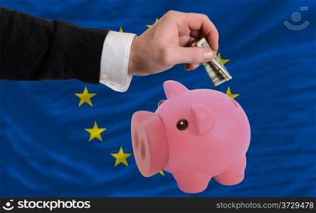 Man putting dollar into piggy rich bank national flag of europe in foreign currency because of inflation
