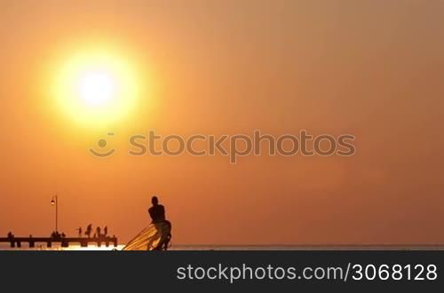 Man raising up his windsurf or sailboard at sunset on a calm ocean against a spectacular vivid orange sky