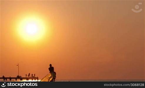 Man raising up his windsurf or sailboard at sunset on a calm ocean against a spectacular vivid orange sky
