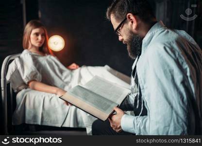 Man reading a book against ill loved woman in hospital bed. Illness of female patient in clinic room, health recovery and treatment. Man reading book against ill woman in hospital bed
