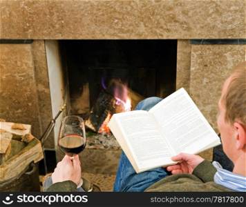 Man reading a book and drinking a glass of wine by the fireplace, relaxing.
