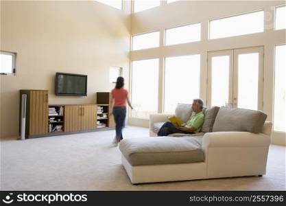 Man reading book in living room with woman walking by