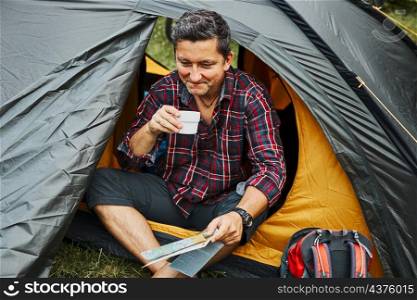 Man relaxing in tent at camping during summer vacation. Man holding map while planning next trip. Actively spending vacations outdoors close to nature. Concept of camp life