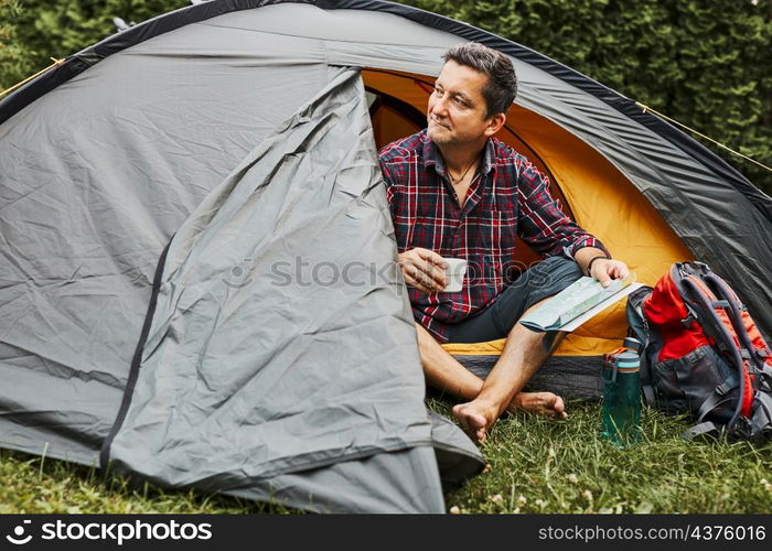 Man relaxing in tent at camping during summer vacation. Man holding map while planning next trip. Actively spending vacations outdoors close to nature. Concept of camp life