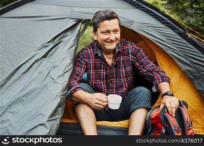 Man relaxing in tent at camping during summer vacation. Relaxing with cup of coffee while sitting at camping. Actively spending vacations outdoors close to nature