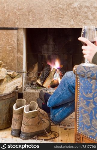 Man relaxing with a glass of red wine by the fireplace after a long hike; his boots off, next to him, warming up