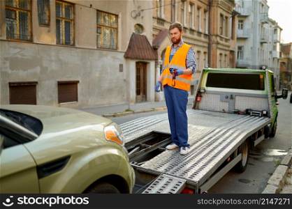 Man road worker in uniform standing on tow truck platform leading broken car loading process. Man road worker leading car evacuation process