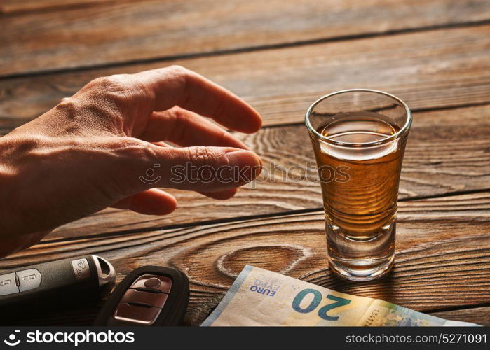 Man&rsquo;s hand reaching to glass of tequila or alcohol drink and car key on rustic wooden table. Drink and drive and alcoholism concept. Safe and responsible driving concept.