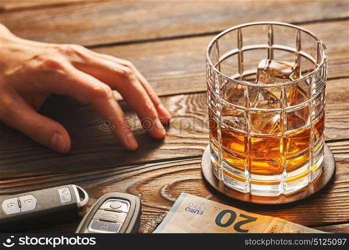 Man&rsquo;s hand reaching to glass of whiskey or alcohol drink with ice cubes and car key on rustic wooden table. Drink and drive and alcoholism concept. Safe and responsible driving concept.