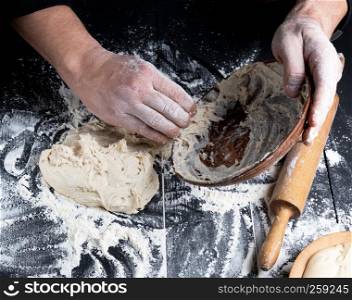 man's hands knead white wheat flour dough on black wooden background