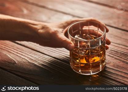 Man's hand holding Glass of whiskey with ice cubes on rustic wooden table with copy-space