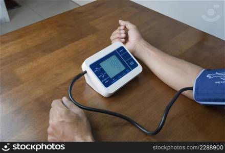 Man's hand using digital sphygmomanometer machine to measuring blood pressure himself on wooden table at home, medical and health care concept