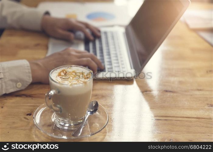 man's hand working with laptop computer with cup of hot latte coffee on wooden table, selective focus and vintage tone