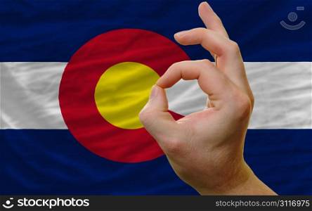 man showing excellence or ok gesture in front of complete wavy american state flag of colorado symbolizing best quality, positivity and succes