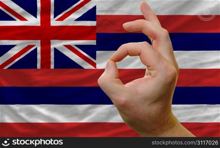 man showing excellence or ok gesture in front of complete wavy american state flag of hawaii symbolizing best quality, positivity and succes