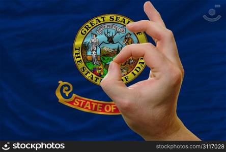 man showing excellence or ok gesture in front of complete wavy american state flag of idaho symbolizing best quality, positivity and succes