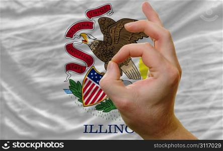 man showing excellence or ok gesture in front of complete wavy american state flag of illinois symbolizing best quality, positivity and succes