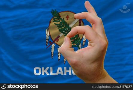 man showing excellence or ok gesture in front of complete wavy american state flag of oklahoma symbolizing best quality, positivity and succes
