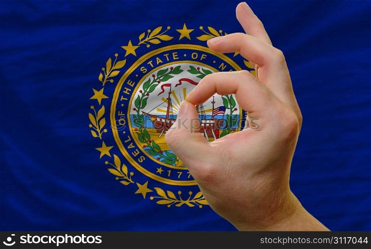 man showing excellence or ok gesture in front of complete wavy american state flag of new hampshire symbolizing best quality, positivity and succes