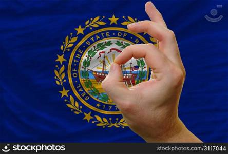 man showing excellence or ok gesture in front of complete wavy american state flag of new hampshire symbolizing best quality, positivity and succes