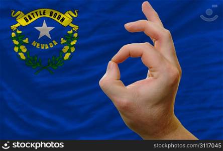 man showing excellence or ok gesture in front of complete wavy american state flag of nevada symbolizing best quality, positivity and succes