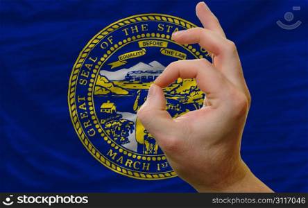 man showing excellence or ok gesture in front of complete wavy american state flag of nebraska symbolizing best quality, positivity and succes