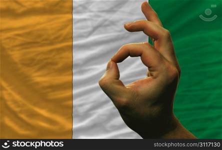 man showing excellence or ok gesture in front of complete wavy ivory coast national flag of symbolizing best quality, positivity and succes