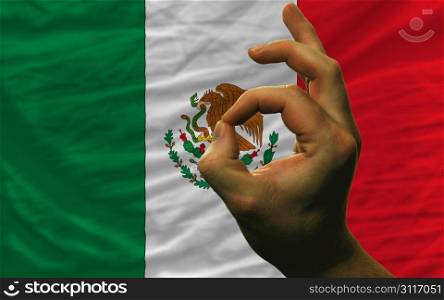 man showing excellence or ok gesture in front of complete wavy mexico national flag of symbolizing best quality, positivity and succes