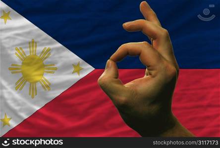 man showing excellence or ok gesture in front of complete wavy philippines national flag symbolizing best quality, positivity and succes