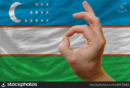 man showing excellence or ok gesture in front of complete wavy uzbekistan national flag of symbolizing best quality, positivity and succes