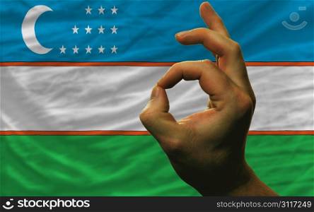 man showing excellence or ok gesture in front of complete wavy uzbekistan national flag of symbolizing best quality, positivity and succes
