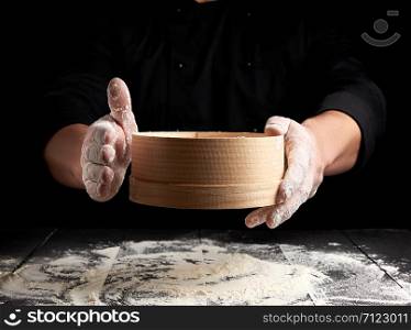 man sifts white wheat flour through a wooden sieve, black background