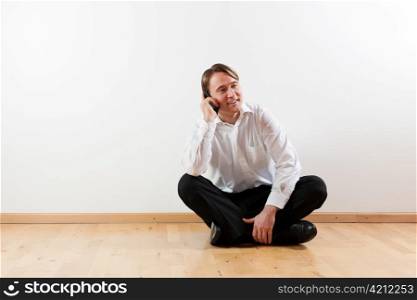 Man sitting cross-legged on the wooden floor of his apartment and is making a phone call