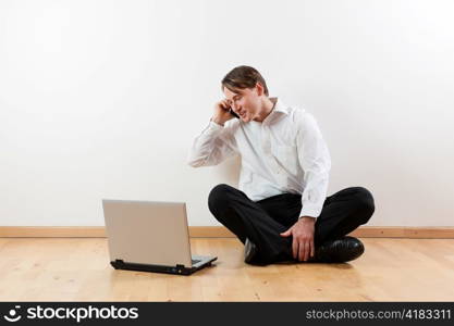 Man sitting cross-legged on the wooden floor of his apartment with laptop and is making a phone call