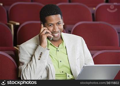 Man sitting in auditorium, using mobile phone and laptop, portrait