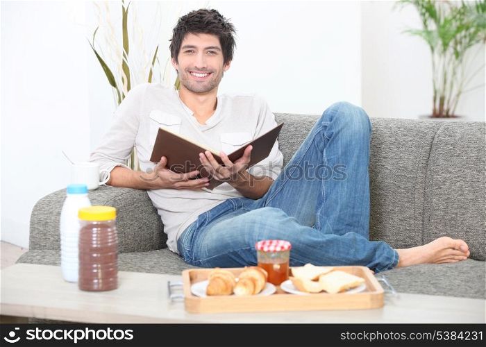 Man sitting on a sofa eating breakfast and reading a book