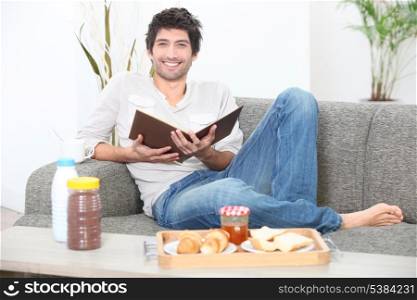 Man sitting on a sofa eating breakfast and reading a book