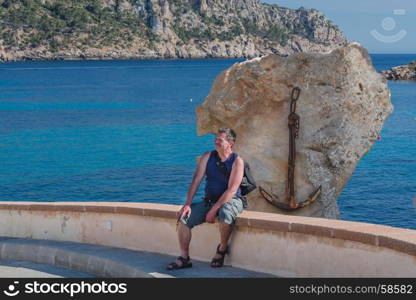 Man sitting on a stone wall facing sideways of the sea and relaxing. Concept of freedom. In the background a rock with a rusty ship anchor. Mallorca, Spain.