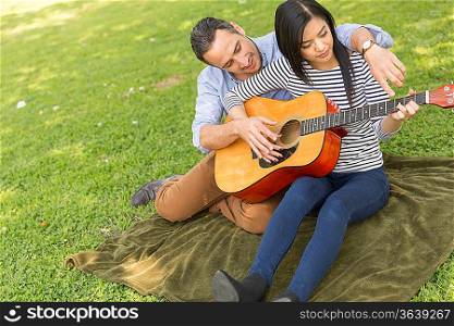Man sitting on grass teaching woman to play acoustic guitar