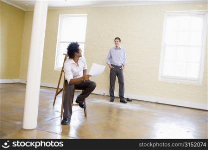 Man sitting on ladder in empty space holding paper talking to other man