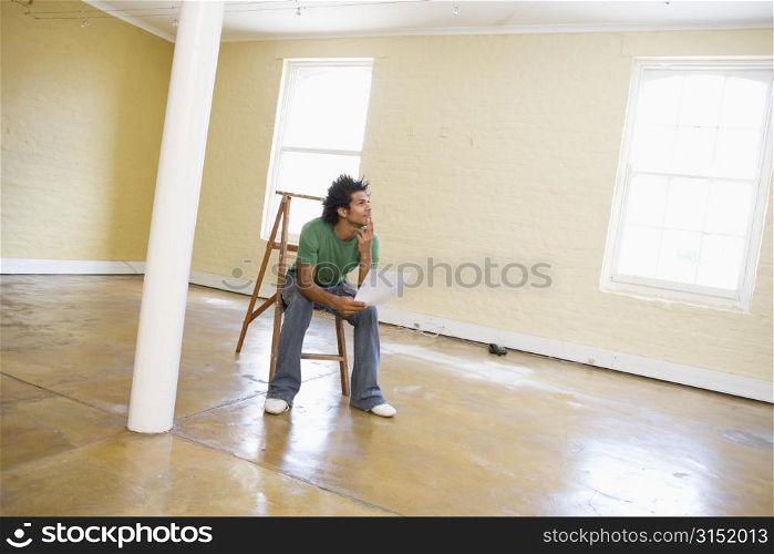 Man sitting on ladder in empty space holding paper thinking