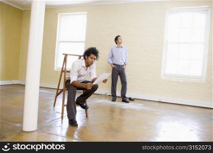 Man sitting on ladder in empty space holding paper with other man behind him