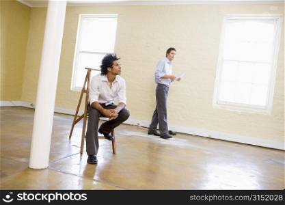 Man sitting on ladder in empty space with another man holding paper