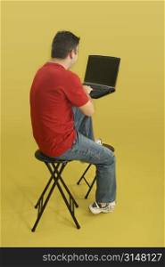 Man sitting on stool with laptop computer. Back side, full body, over yellow.