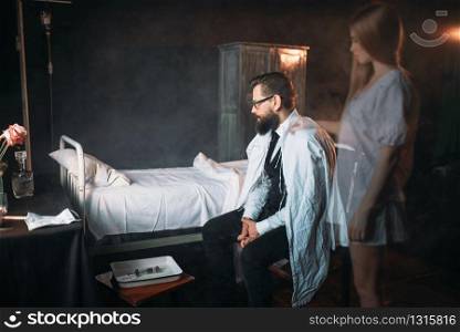 Man sitting on the chair against empty hospital bed, soul of the dead loved woman standing behind. Death of female patient in clinic. Man against empty hospital bed, soul of dead woman