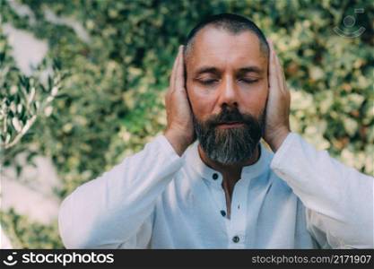 Man sitting with his eyes closed and doing a Reiki auto treatment, holding hands on ears. . Reiki Self-Treatment.