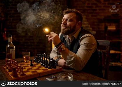Man smokes a cigar, drinks alcohol beverage and play chess, bookshelf and vintage office interior on background. Tobacco smoking culture, specific flavor. Male smoker leisures at the chessboard. Man smokes a cigar, smoker leisures at chessboard