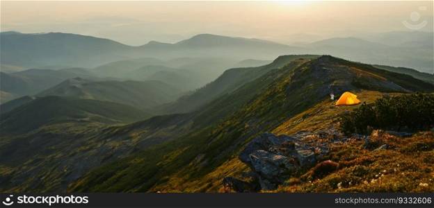 Man spending summer vacation in mountains, standing by tent looking at sunrise over valley. Active man hiking in mountains. Yellow tent put over valley. Foggy mountain landscape view at sunrise