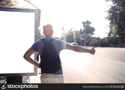 Man standing alongside a blank billboard on a bus stop on a street thumbing a lift backlit by a bright summer sun