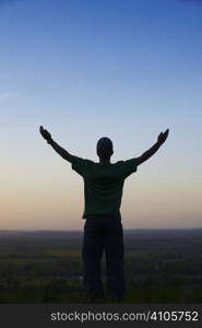 Man standing with arms outstretched in front of landscape at dusk